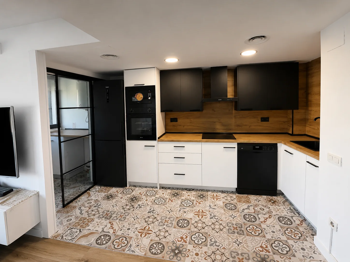 Cocina con muebles blancos y negros, frente de madera y pavimento de baldosas decorativas.